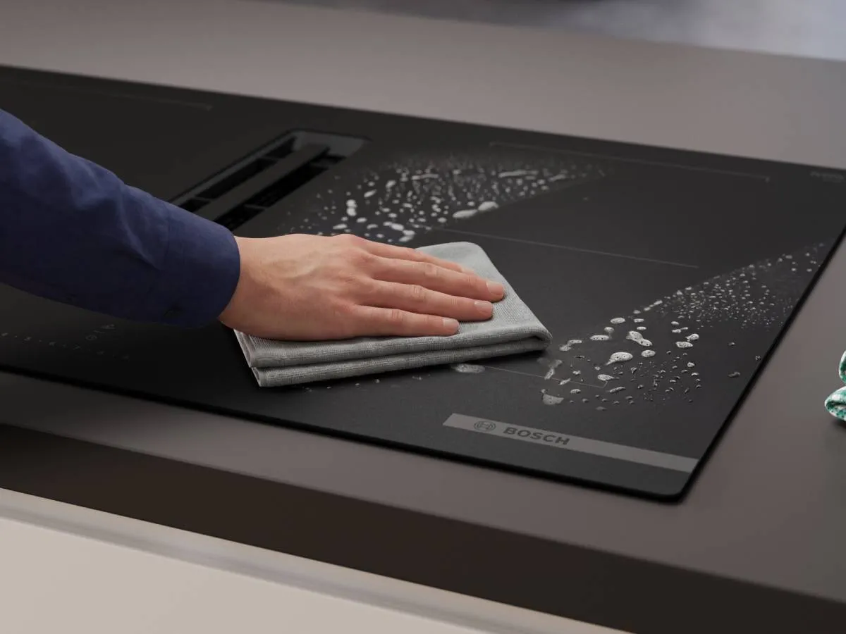 A hand wipes down a black Bosch induction cooktop with an integrated downdraft extractor, showing water droplets and suds, highlighting easy cleaning.