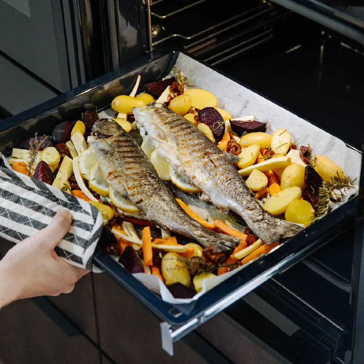 Zwei ganze gegrillte Fische auf Zitronenscheiben mit gemischtem geröstetem Gemüse werden auf einem Backblech aus dem Ofen genommen.