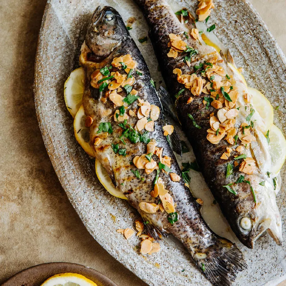 Zwei ganze gegrillte Fische, garniert mit Mandelscheiben und gehackten Kräutern, serviert auf Zitronenscheiben auf einer rustikalen ovalen Platte.