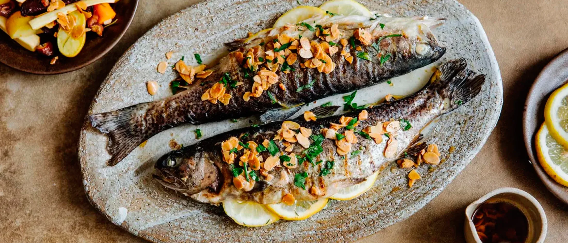 Zwei ganze gegrillte Fische, garniert mit Mandelscheiben und gehackten Kräutern, serviert auf Zitronenscheiben auf einer rustikalen ovalen Platte.