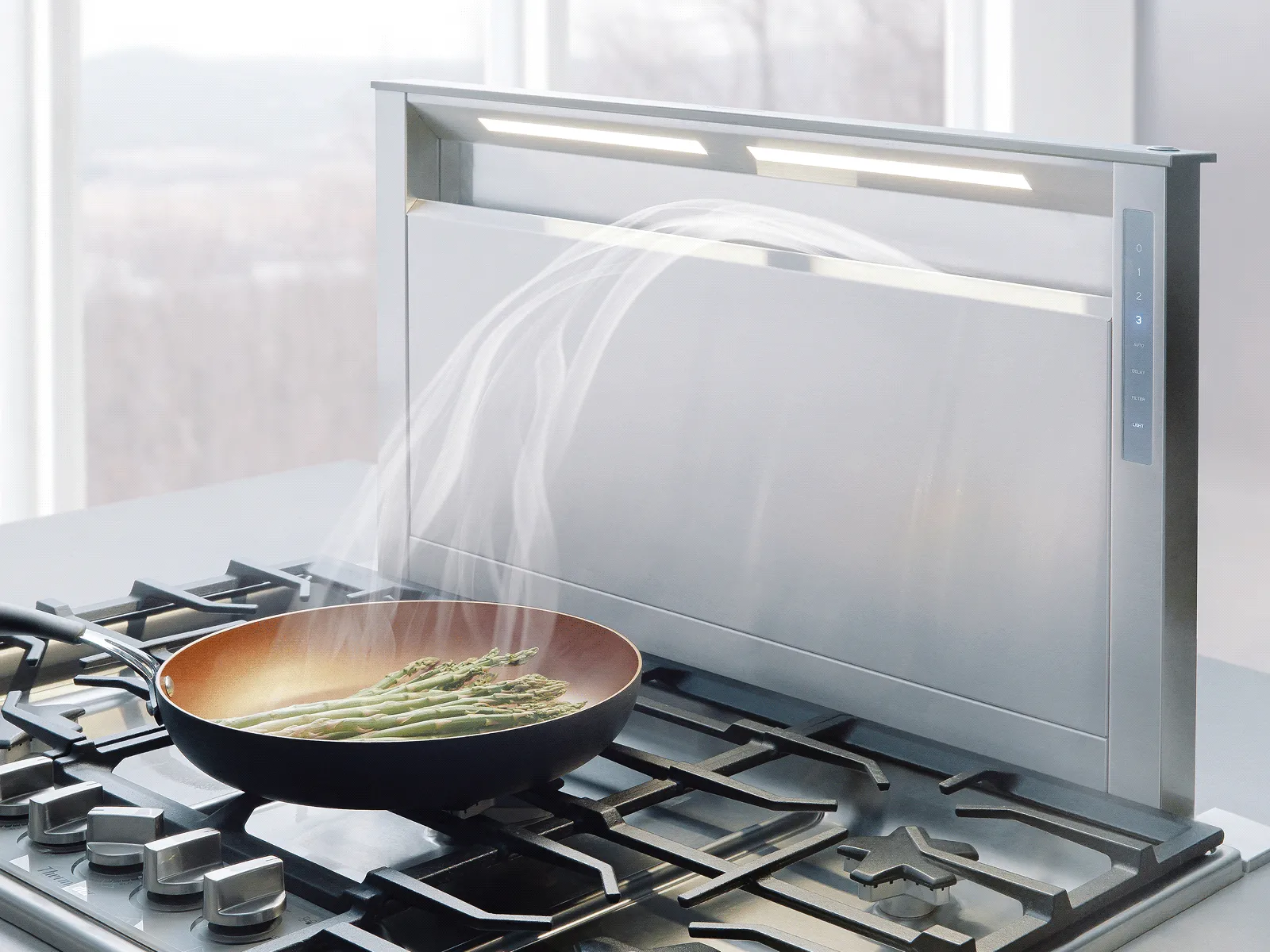 Modern kitchen cooktop with a telescopic downdraft ventilation system extracting steam from a pan of asparagus, illuminated by integrated lighting.