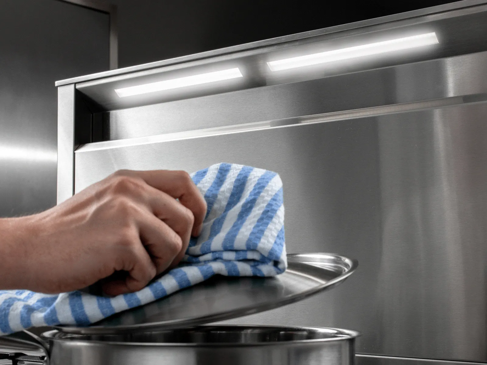 Close-up of a hand lifting a pot lid with a cloth over a cooktop, illuminated by the integrated LED lights of a stainless steel telescopic downdraft ventilation system.