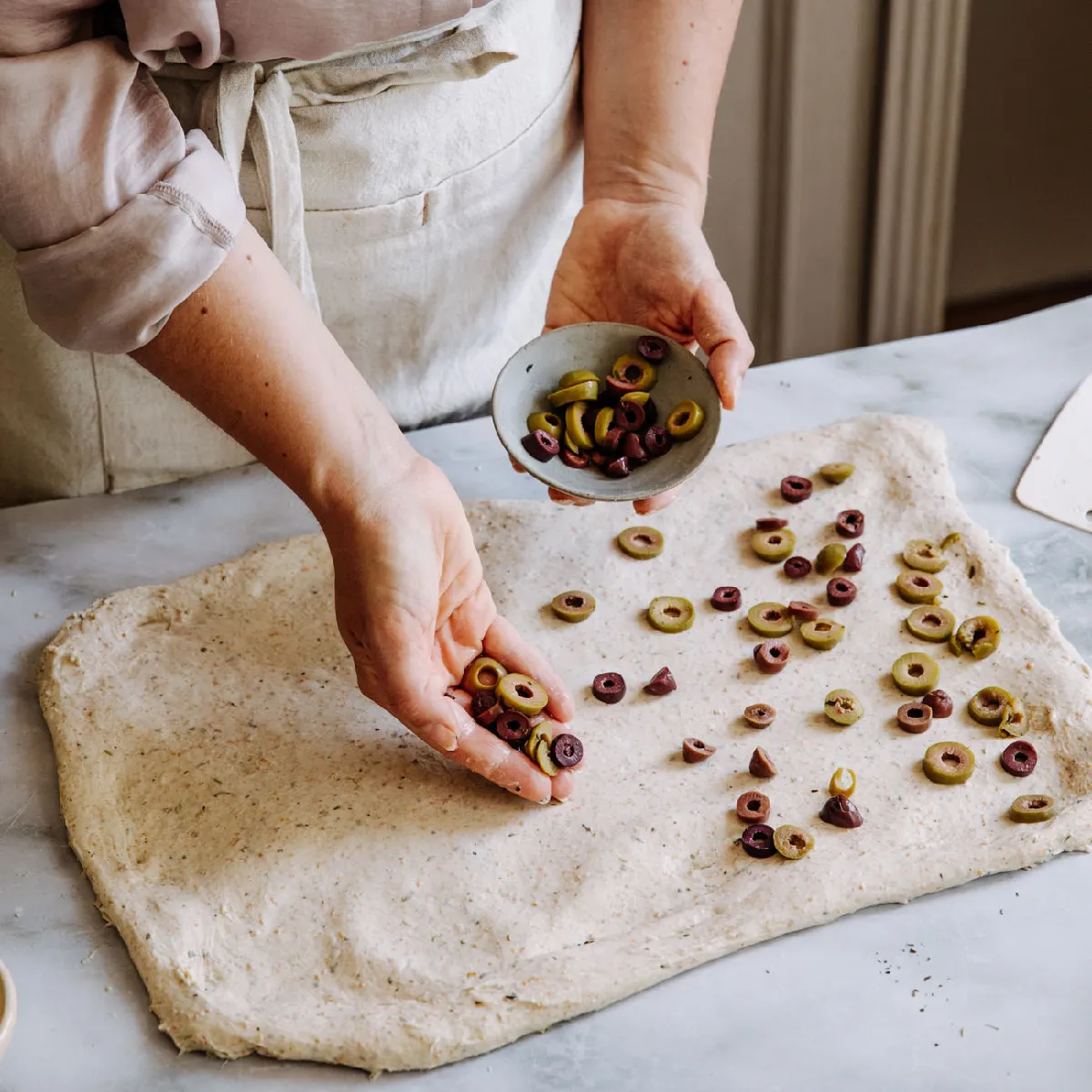 Personne saupoudrant des olives vertes et noires tranchées sur une pâte étalée sur une surface en marbre, en train de préparer de la nourriture.