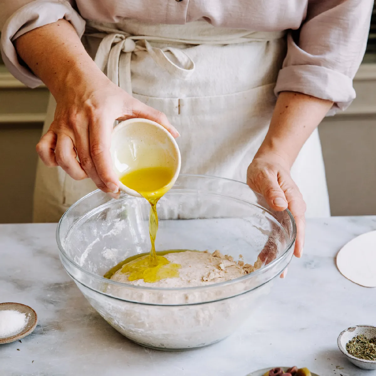 Personne versant de l'huile d'olive dans un bol en verre avec de la pâte à base de farine sur un comptoir en marbre, portant un tablier.