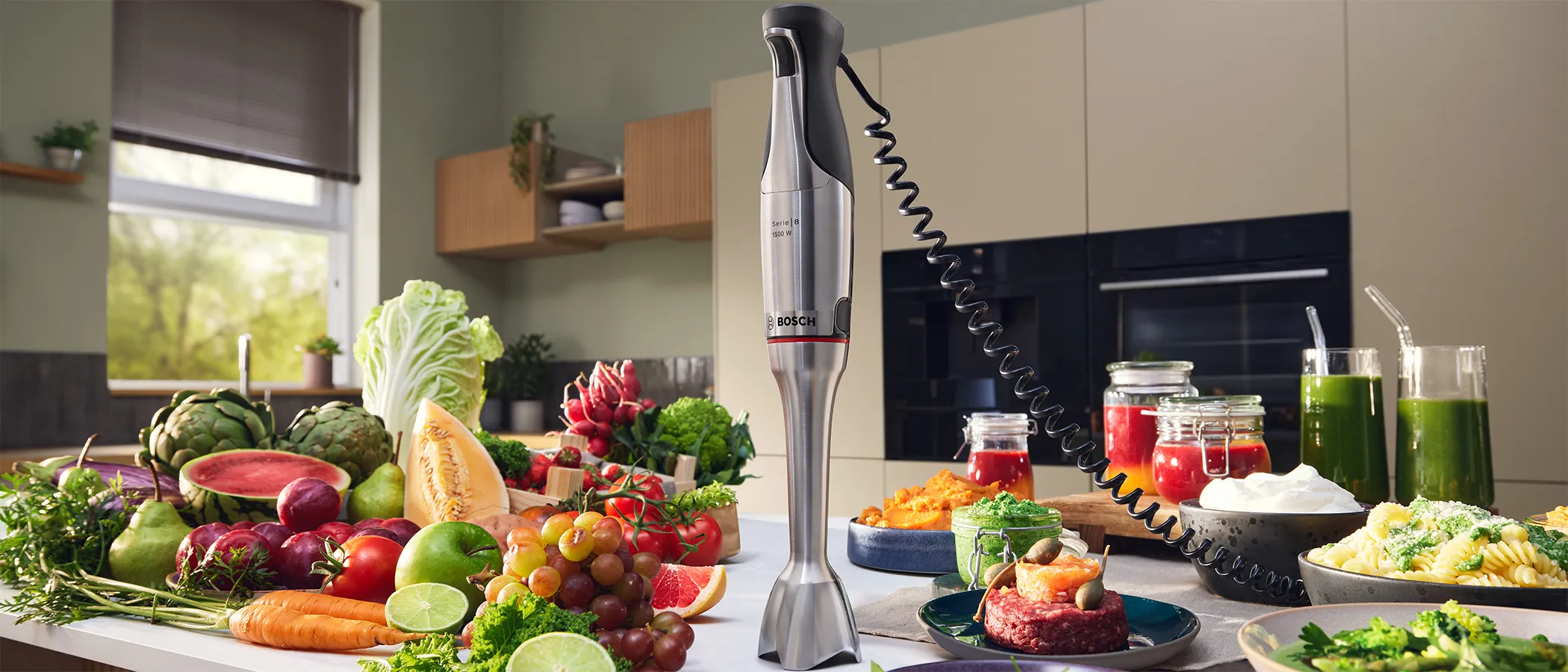 Uśmiechnięta kobieta miksuje zielone smoothie w jasnej kuchni, świeże liście fruwają wokół niej, a w jej otoczeniu są orzechy, ser i bazylia.