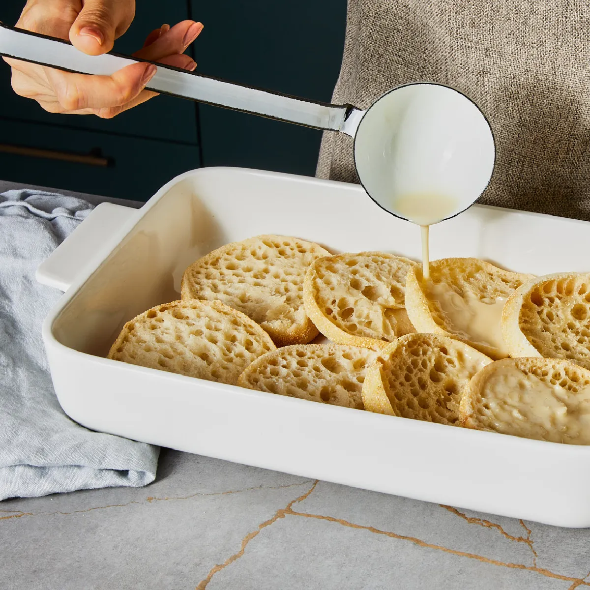 Baked bread slices in a white dish, with a hand pouring a liquid over them, on a gray background.
