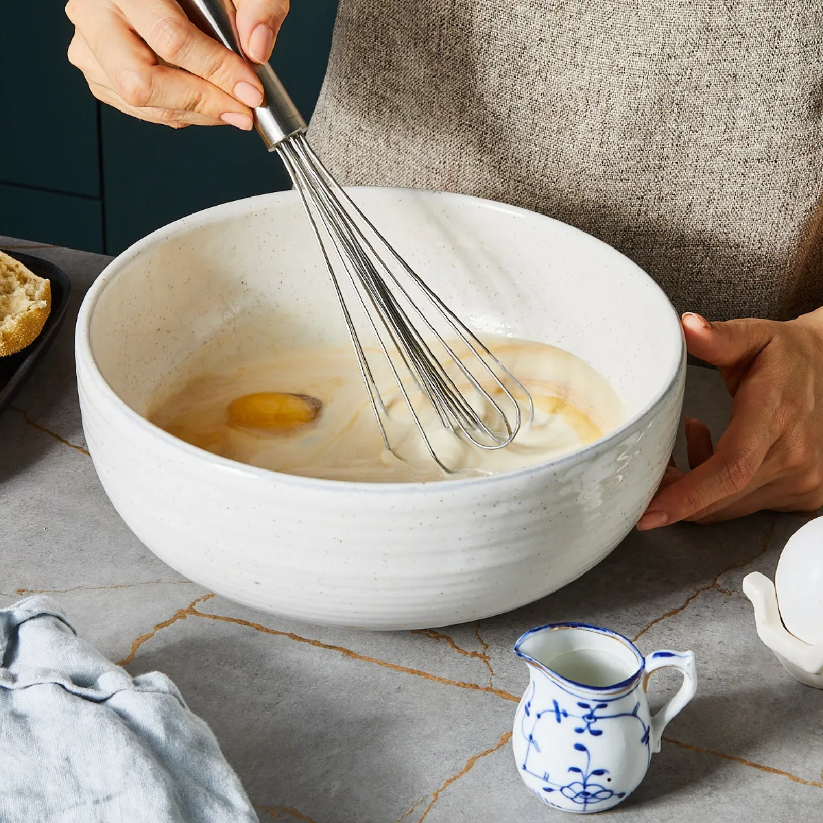 Hands whisking eggs in a bowl, surrounded by baking ingredients like bread, eggs, and a small pitcher.
