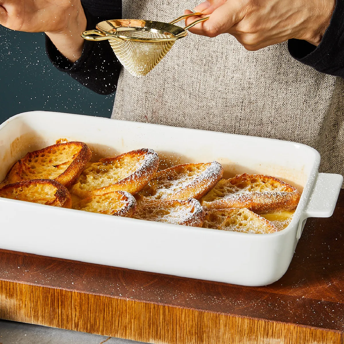 Baked bread slices in a white baking dish, being sprinkled with powdered sugar by a person's hands.