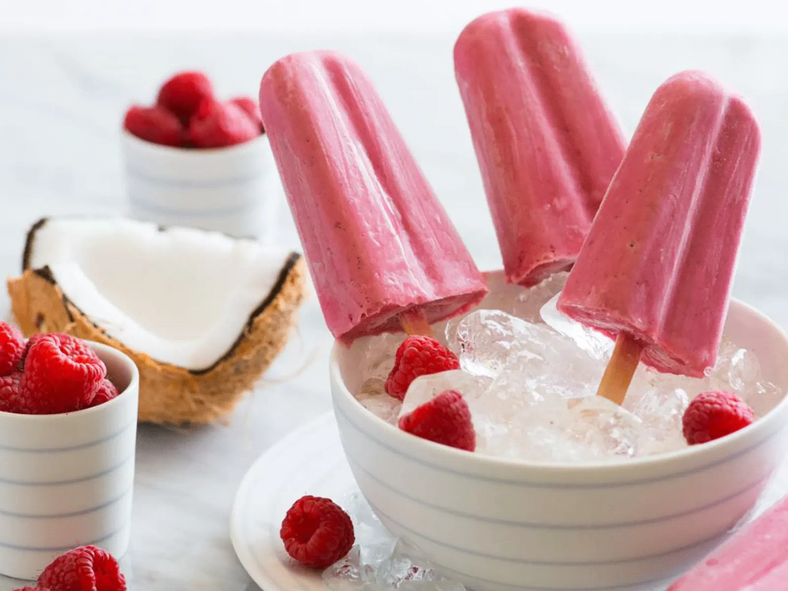 Three pink raspberry-coconut popsicles standing in a bowl of crushed ice, surrounded by scattered raspberries, a halved coconut showing the white flesh