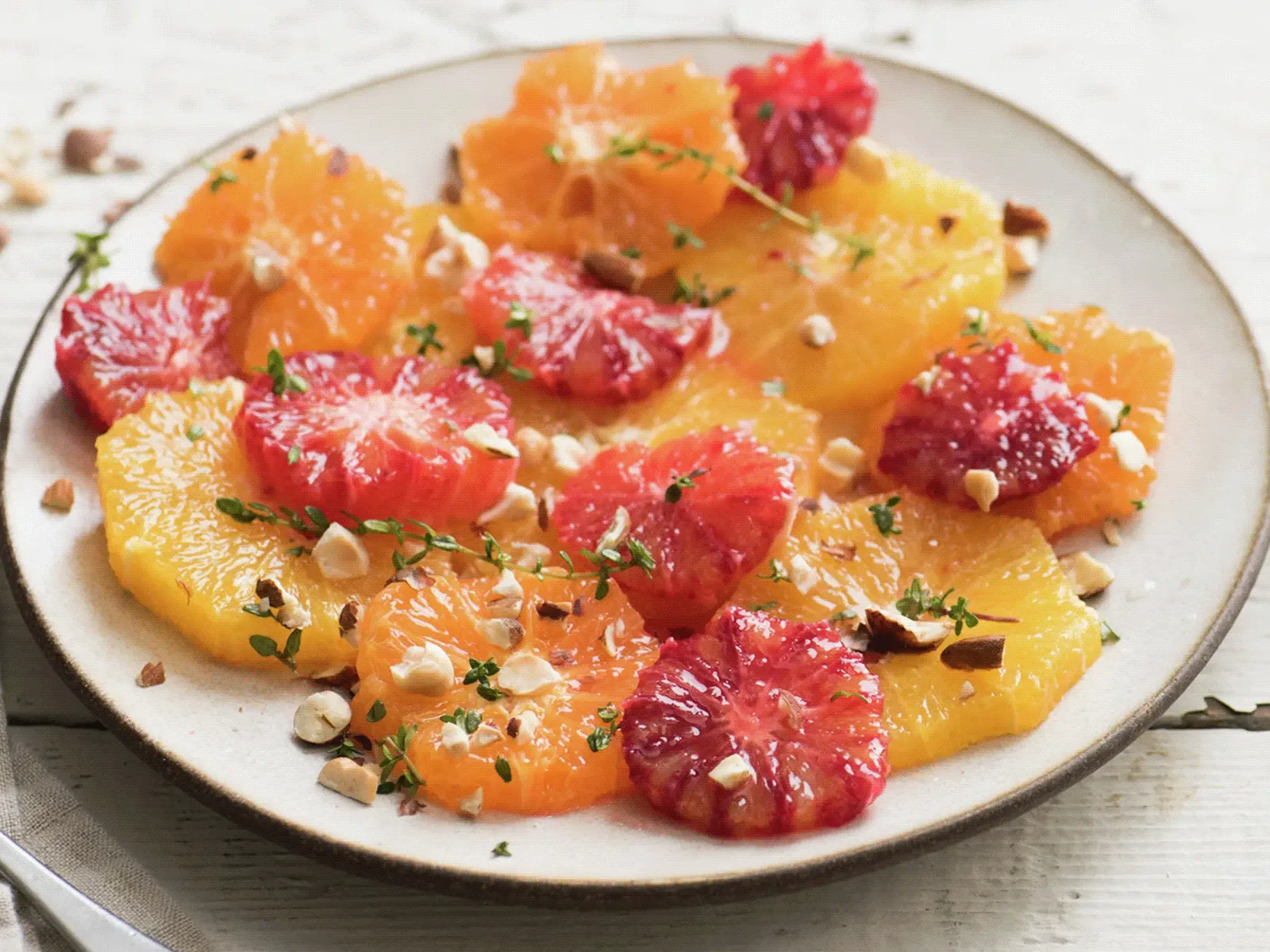 Top-down view of a plate of citrus salad with alternating red and orange citrus slices, dotted with small mounds of ruby-red pulp and sprinkled with chopped nuts and fresh thyme leaves, set on a light table with small bowls at the edges.