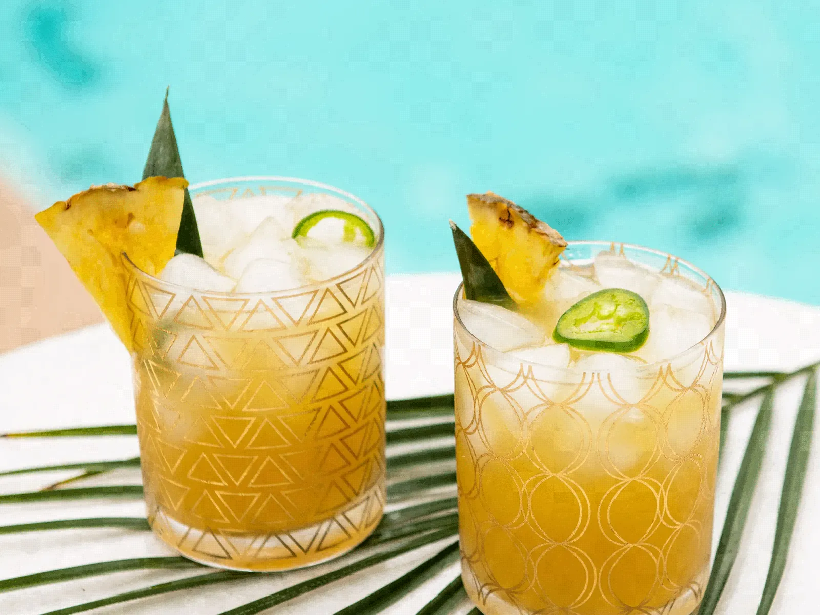 Two pineapple jalapeño cocktails garnished with pineapple slices and jalapeño rings, set beside a pool on a white surface.