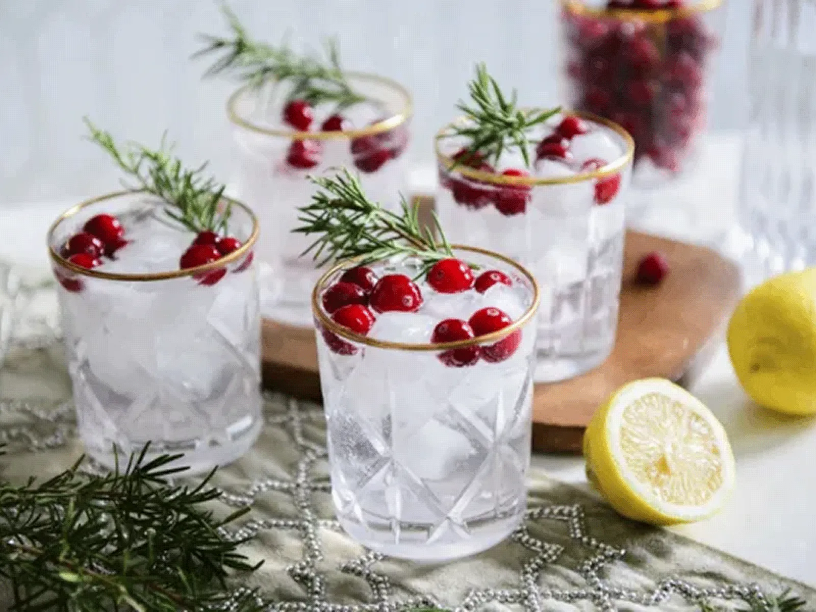 Four clear glasses with ice, red berries, and rosemary sprigs, with a halved lemon and additional greenery on a white surface.