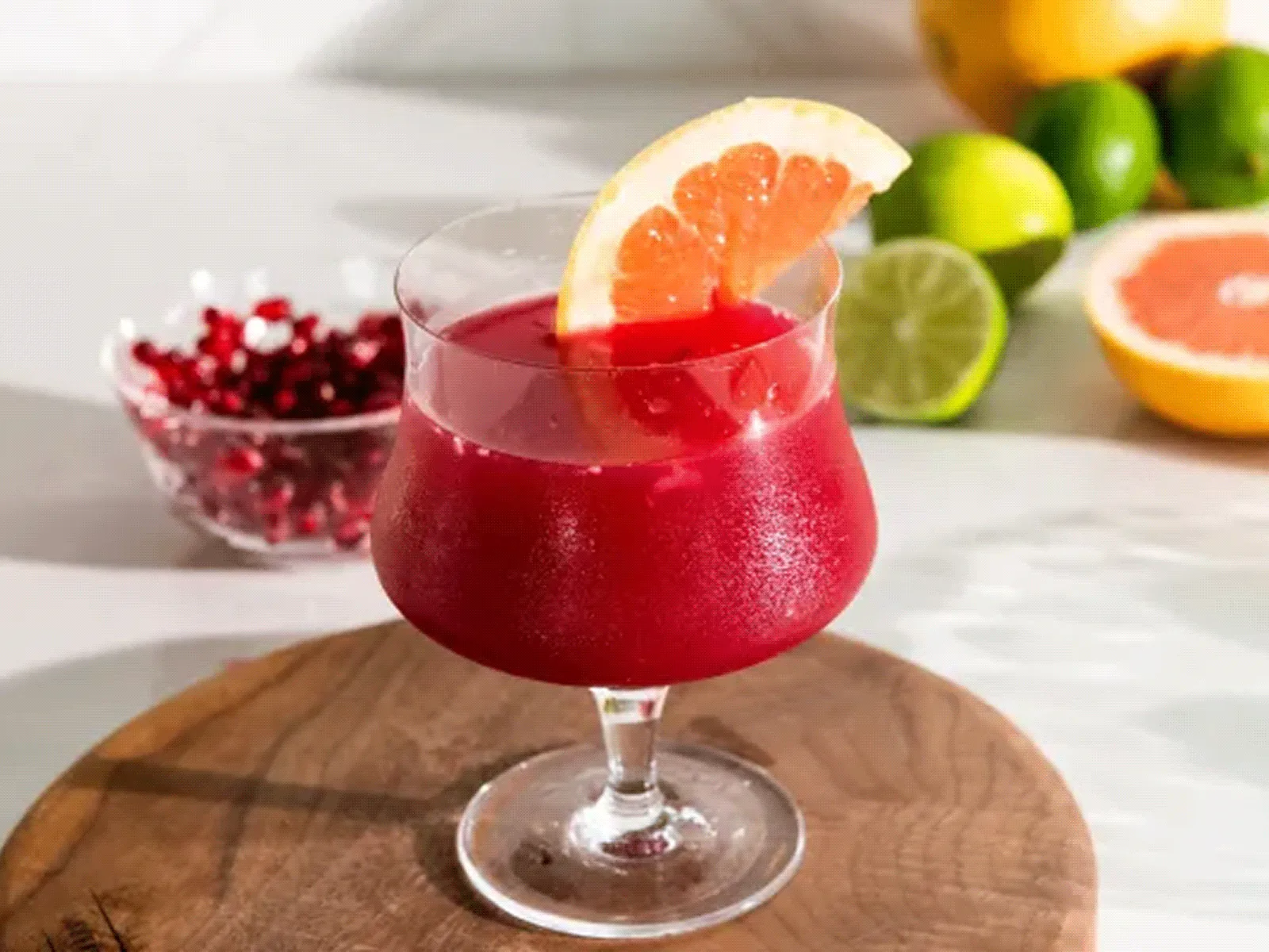 Stemmed cocktail glass on a round wooden board holding a deep red pomegranate-style drink topped with a grapefruit wedge and a few pomegranate arils; a small bowl of pomegranate seeds and halved/whole citrus (limes and grapefruit) sit blurred on a white countertop behind it.