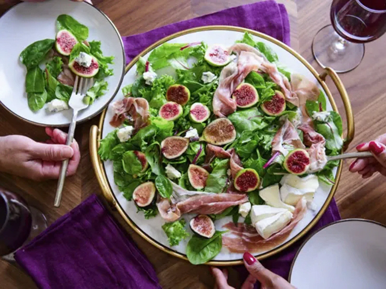 Top-down view of a large round platter of mixed greens topped with halved figs, thin slices of prosciutto and pear slices; three hands reach in with forks and plates on a wooden table with a purple napkin and glasses of red wine.