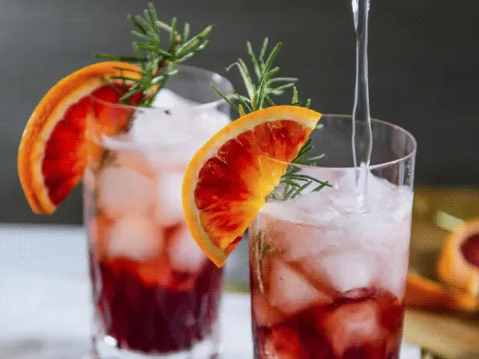 Two tall glasses of iced deep-red cocktails on a light surface, each garnished with a blood orange slice and a rosemary sprig; a clear liquid is being poured into the right glass and a wooden board with orange wedges sits blurred in the background.