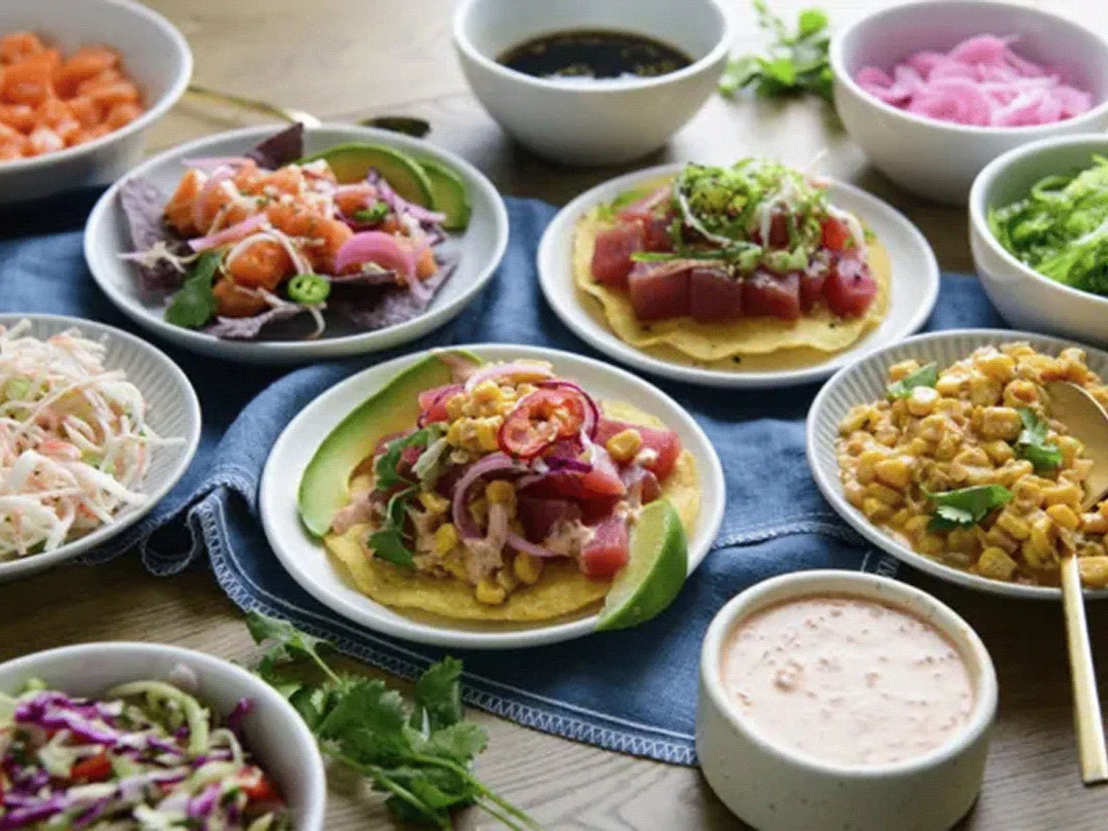 Assorted colorful dishes including ceviche-style seafood on tostadas, surrounded by bowls of sauces, pickled onions, and fresh herbs.