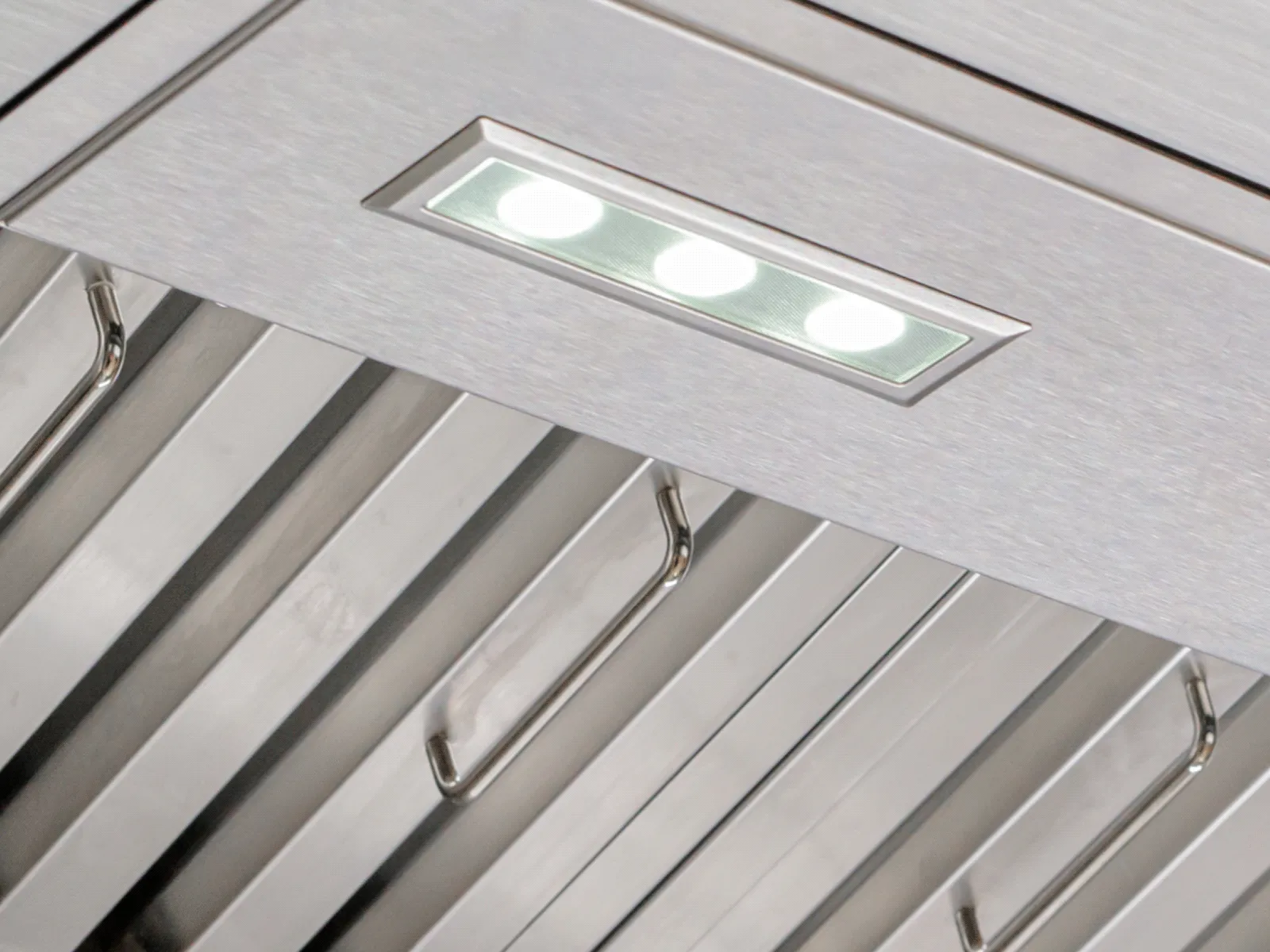 An extreme close-up on a built-in rectangular LED light on a brushed stainless steel range hood. Below the light, the handles of the baffle filters are visible.