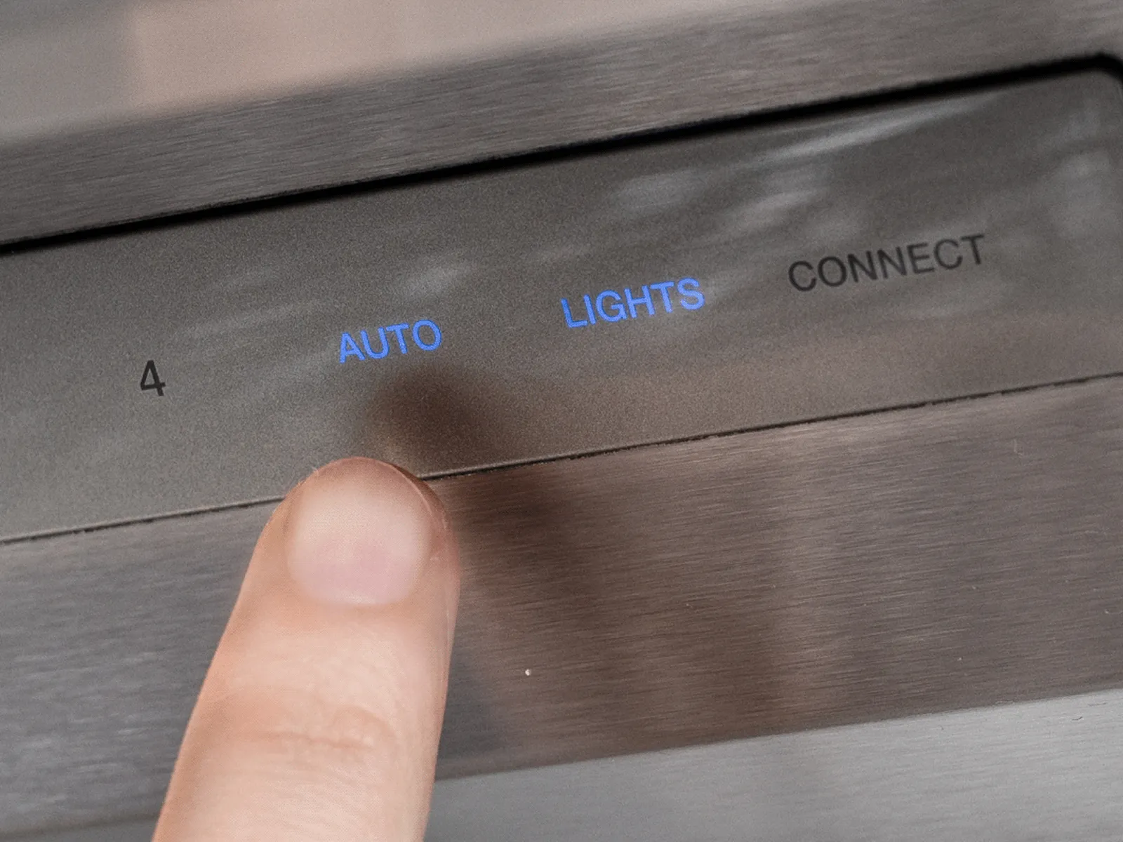 Close-up of a finger pressing the touch-sensitive "AUTO" button on a stainless steel appliance control panel. The button is illuminated in blue, and other controls for "LIGHTS" and "CONNECT" are visible nearby.