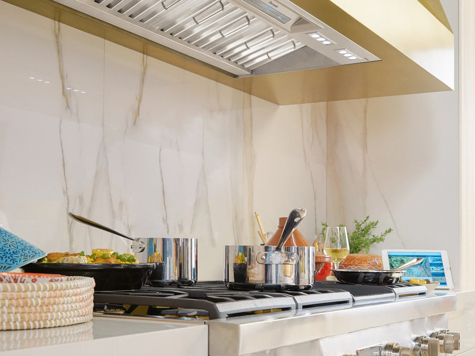 A luxurious kitchen scene featuring a stainless steel cooktop with various pots and pans. A sleek, built-in range hood with a decorative gold-toned trim is mounted above, against a white marble backsplash.