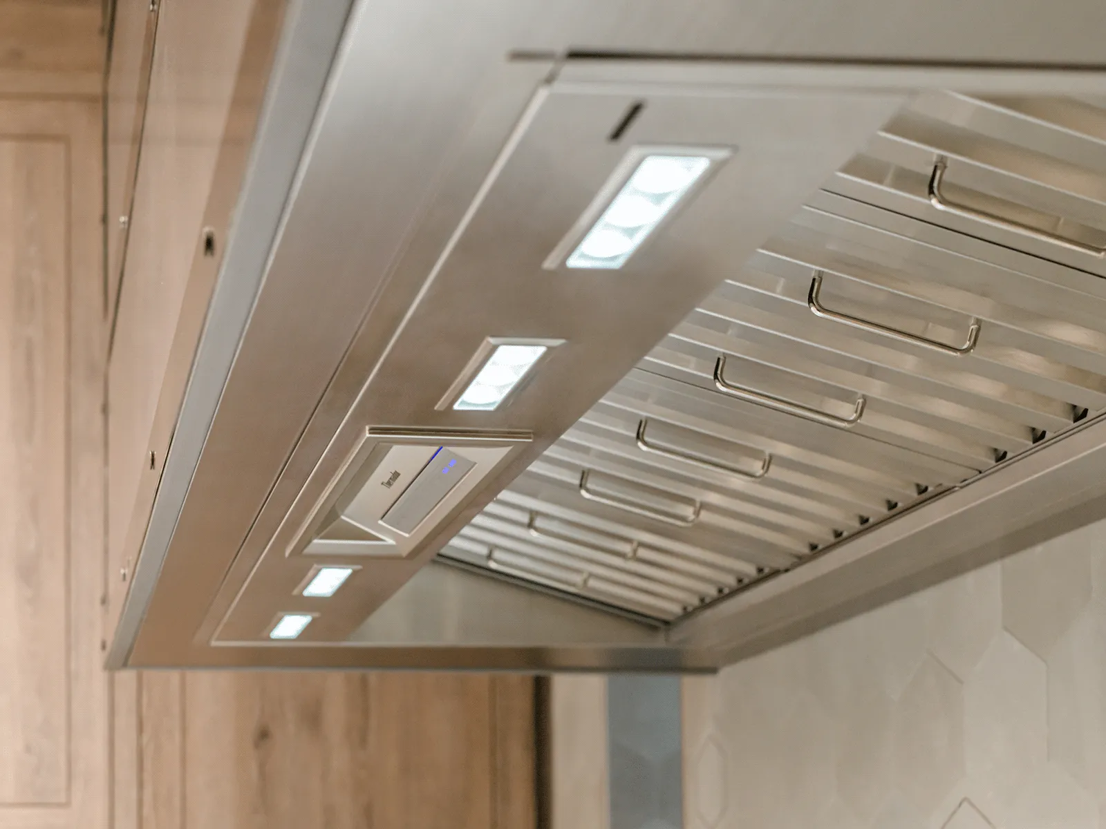 An upward-angle, close-up view of the underside of a stainless steel Thermador range hood. The image highlights the sleek baffle filters and powerful, built-in LED lighting.