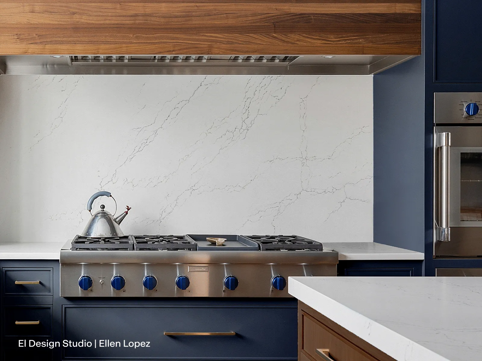 A Thermador Pro Rangetop, featuring its signature blue knobs and a central griddle, is set into a white quartz countertop. The kitchen features dark blue shaker-style cabinets, a full-height white quartz backsplash with grey veining, and a stainless steel ventilation hood framed by a custom wood surround. To the right, a built-in stainless steel wall oven is visible.