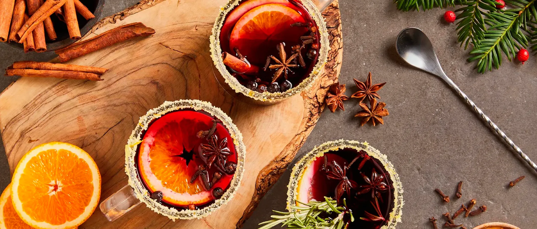 Three glasses of mulled wine garnished with orange slices, star anise, cinnamon, and spices on a wooden board and gray surface.