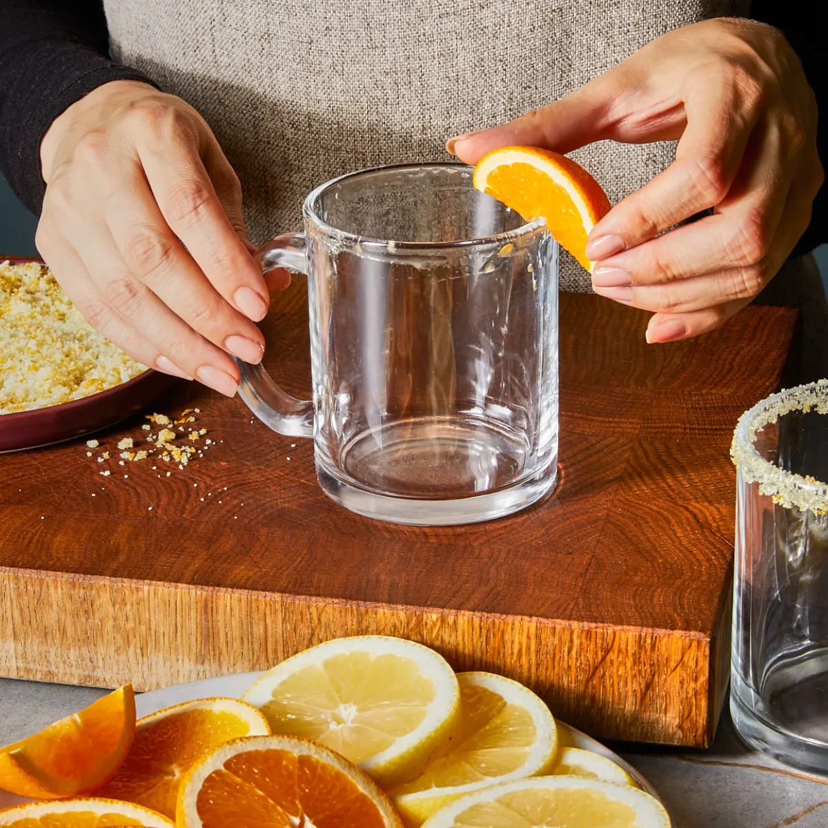Hands garnishing a glass mug with an orange slice on a wooden cutting board, with lemon and orange slices on a plate nearby.