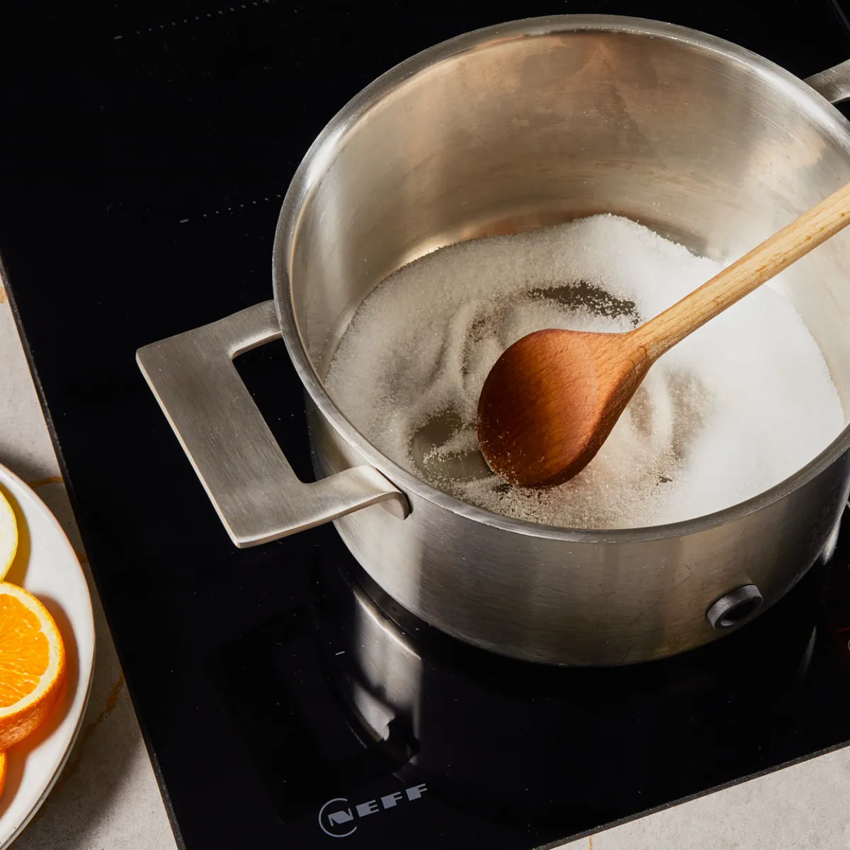 Metal pot with sugar being stirred by wooden spoon on stovetop, with sliced citrus fruit on a plate nearby.