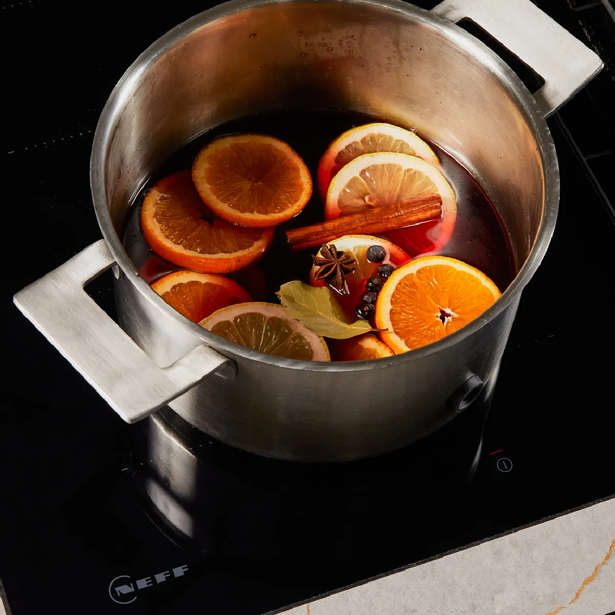 Stainless steel pot on stovetop with simmering mulled wine, orange and lemon slices, cinnamon stick, star anise, bay leaf, and peppercorns.