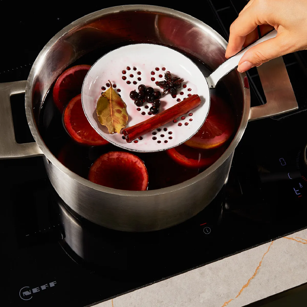 Hand holding a perforated ladle over a pot of simmering spiced mulled wine with orange slices, cinnamon, bay leaf, and star anise.
