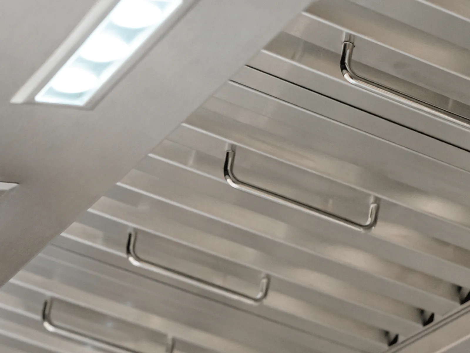 A close-up, angled view of the underside of a stainless steel ventilation hood. The image highlights the professional-style, dishwasher-safe baffle filters with small handles for easy removal. A bright, rectangular LED light illuminates the sleek, high-quality construction of the appliance.