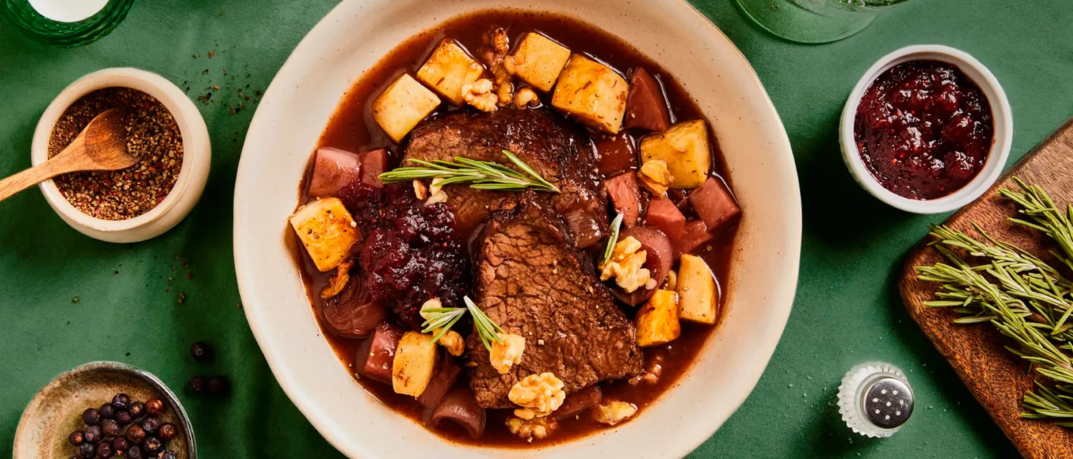 Bowl of beef stew with root vegetables, cranberries, walnuts, and rosemary on a green tablecloth with spices and herbs nearby.
