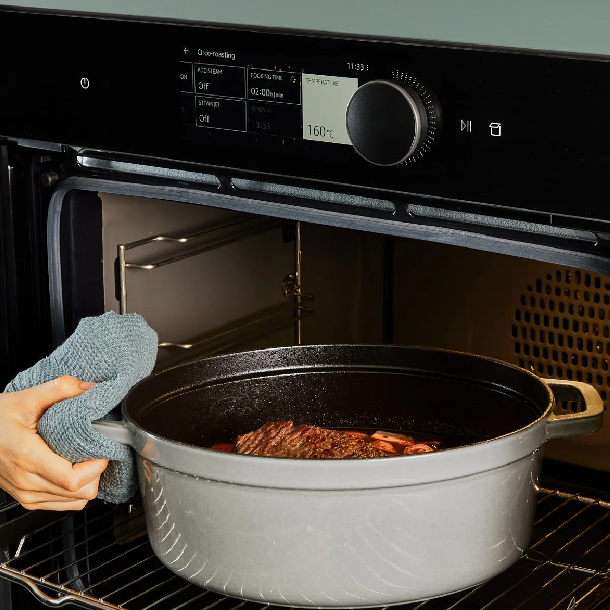 Hand using a cloth to place a cast iron pot with food into a modern built-in oven set at 160°C.
