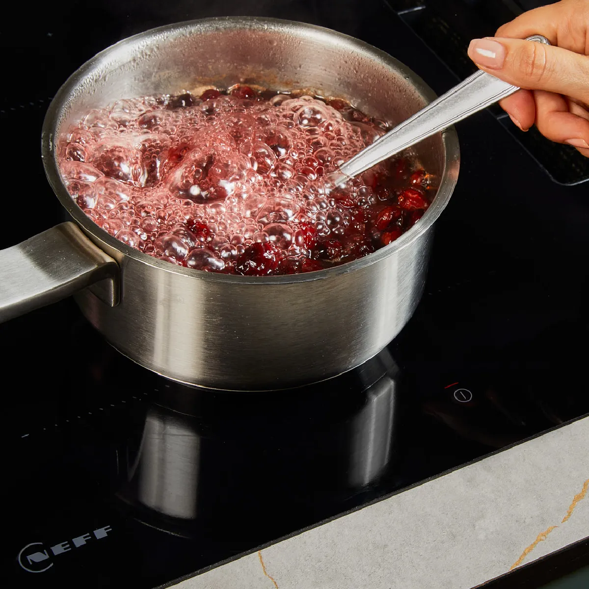 Hand stirring a bubbling red liquid cooking in a stainless steel pot on a black induction stove.