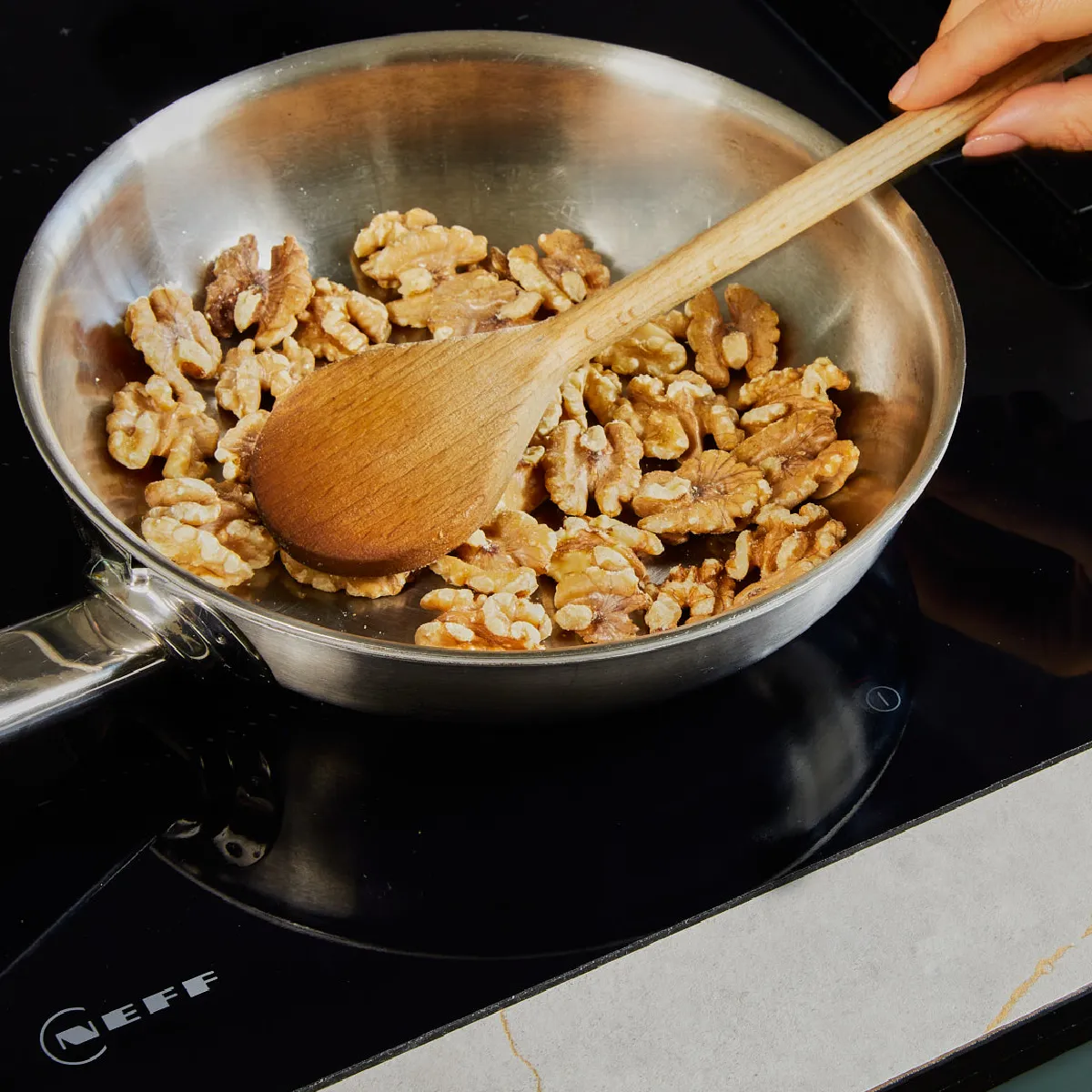 Hand stirring walnuts in a stainless steel pan on a black electric stove with a wooden spoon.