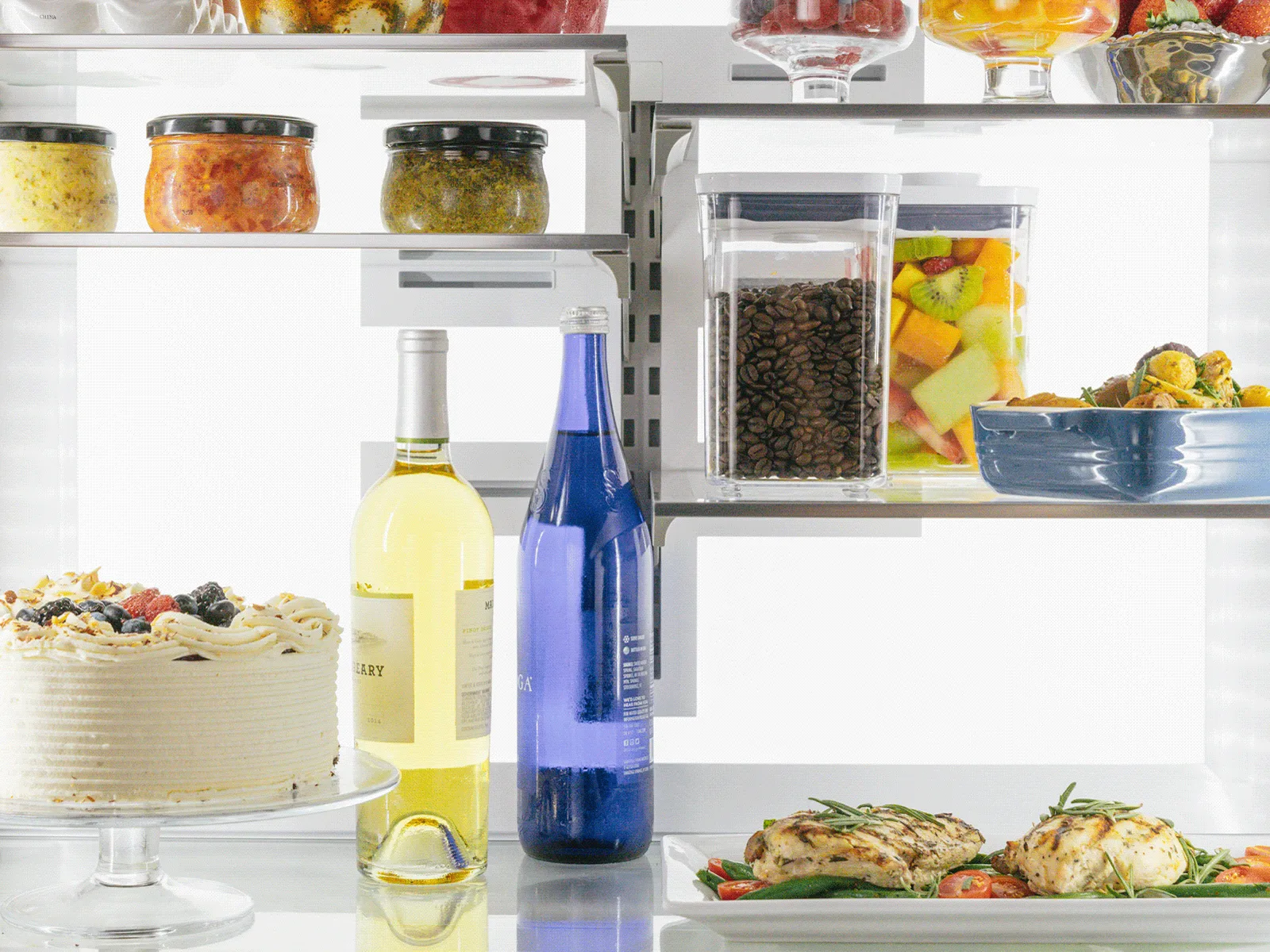 A refrigerator shelf holding a cake, wine, grilled chicken, and containers of fruit and coffee.