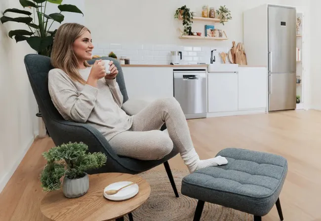 Una mujer vestida con ropa cómoda está sentada en una silla con una taza, con las piernas apoyadas en un puf, en una moderna cocina-sala de estar con plantas y decoración minimalista.