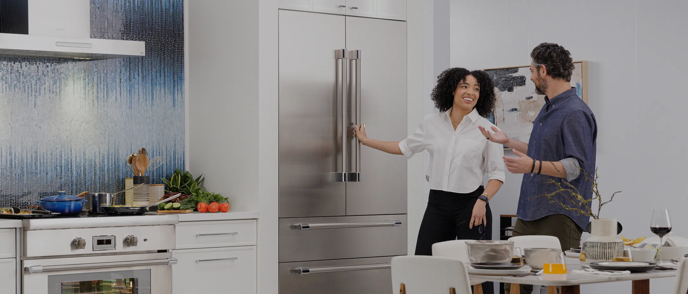 Built-in stainless steel French door refrigerator in a modern kitchen, with two people standing beside it while preparing food on the countertop and a dining table set nearby.