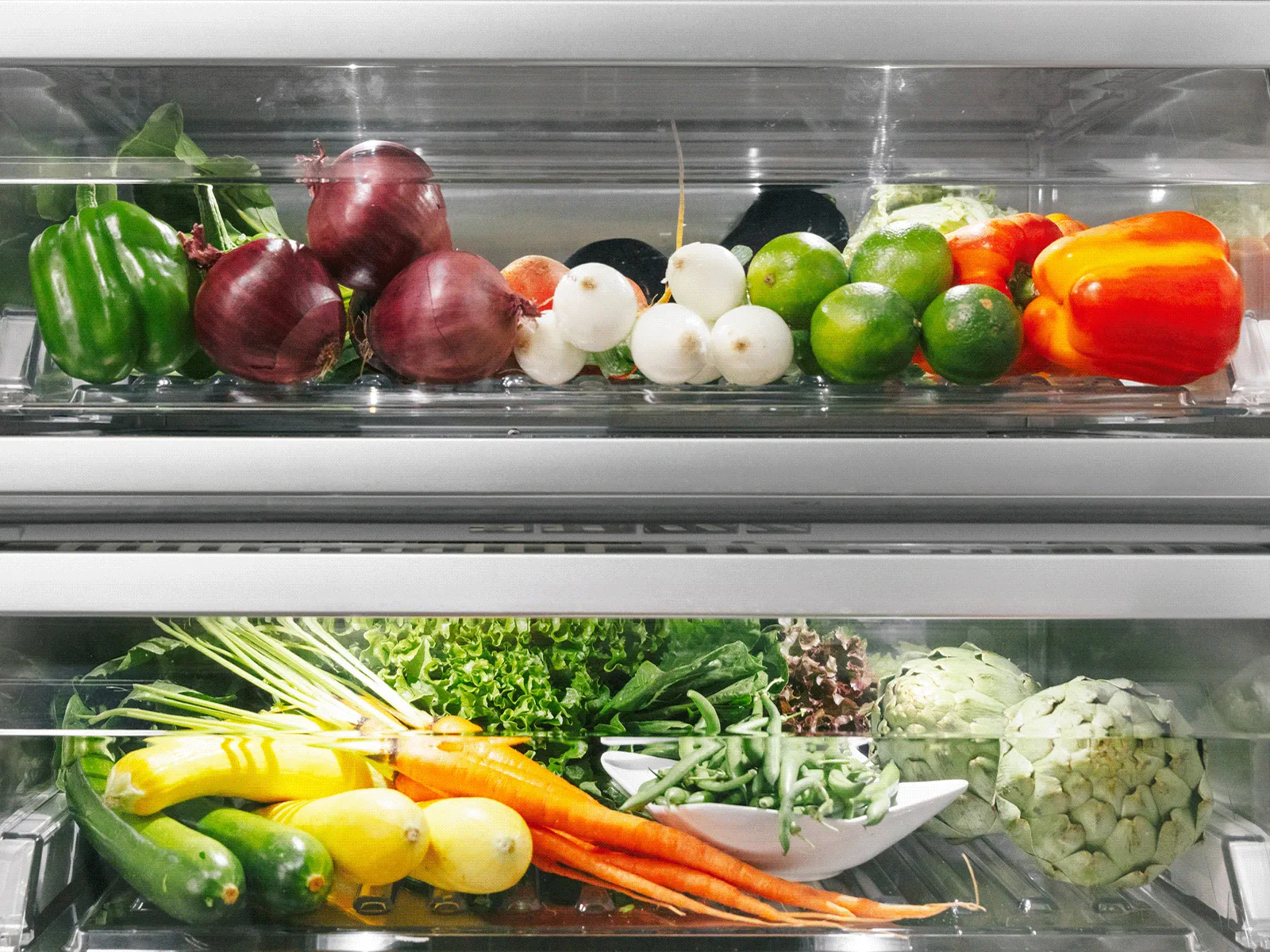 Close-up of two transparent refrigerator crisper drawers filled with fresh vegetables: the top drawer holds green bell peppers, red onions, small white radishes, limes and a red pepper; the bottom drawer contains carrots with tops, yellow squash/zucchini, assorted leafy greens and a head of cauliflower
