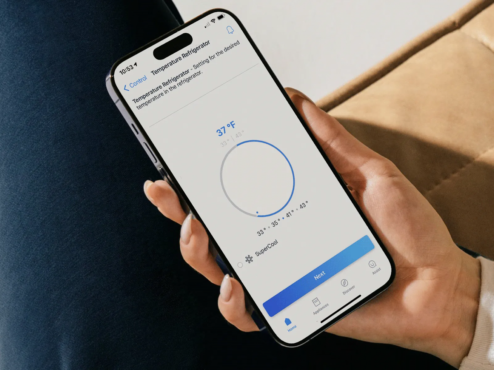 Hand holding a smartphone displaying a temperature-control app with a circular dial set to 37°F and a blue action bar at the bottom; sofa and cushion visible in the background.