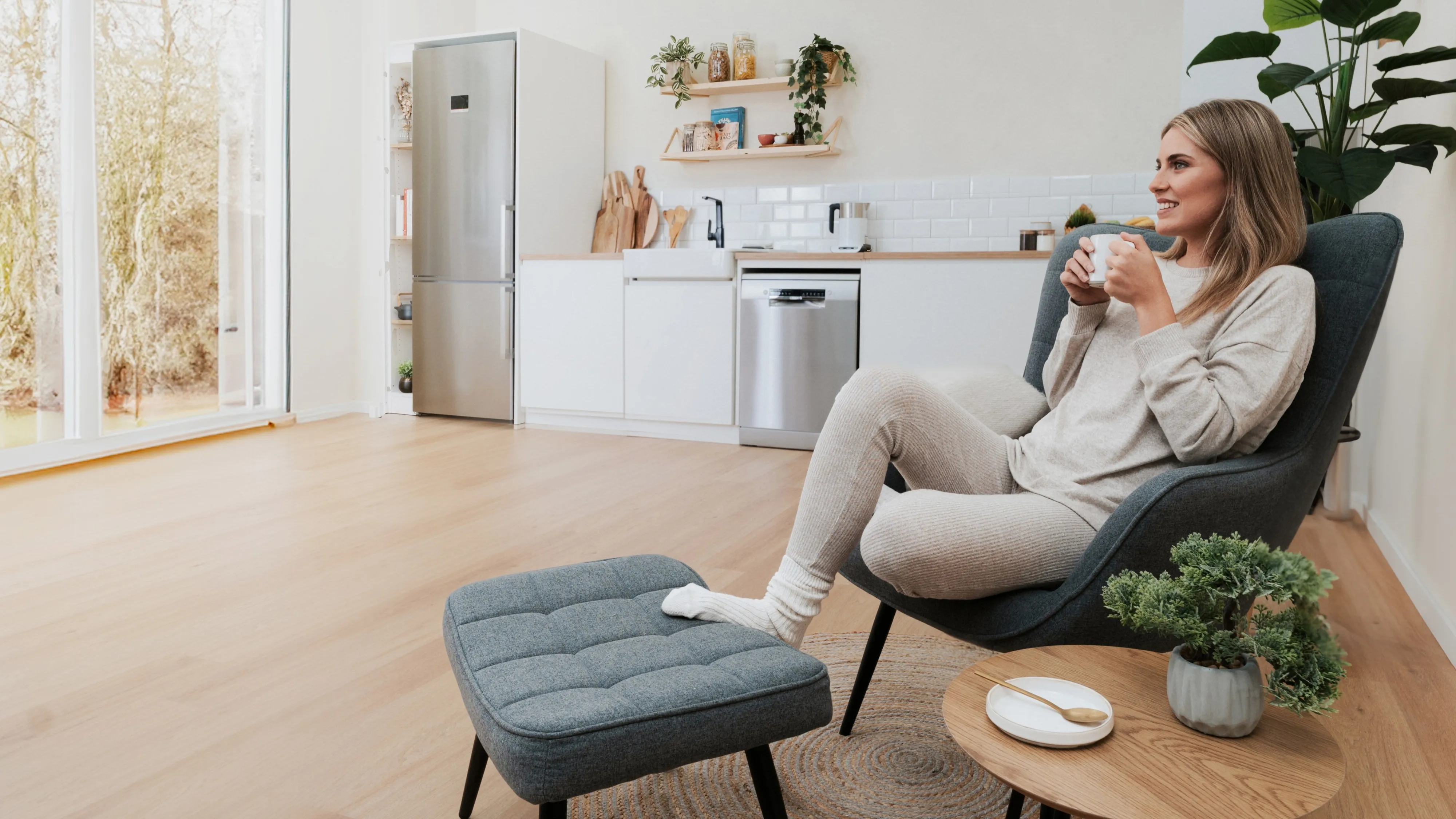 Une fille assise sur une chaise, en train de prendre tranquillement un café, avec en arrière-plan une cuisine dotée d'une grande fenêtre.