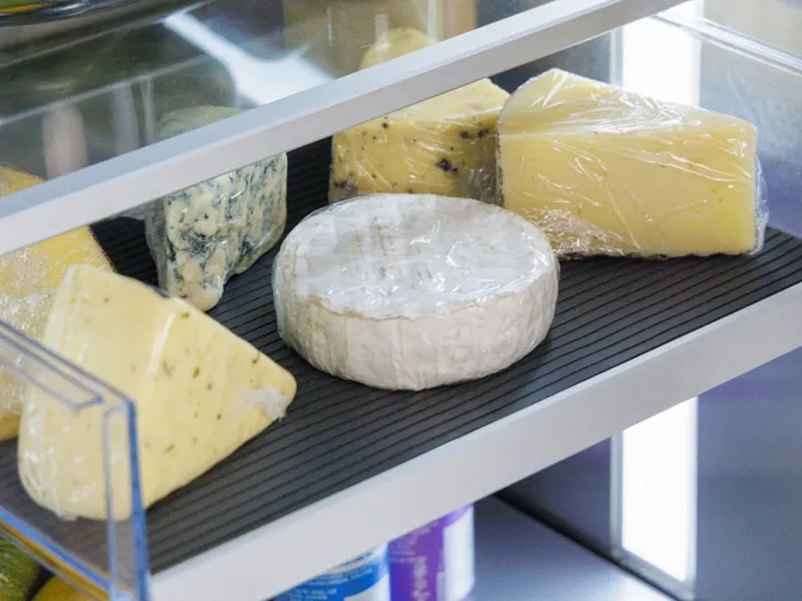 Close-up of various cheeses, including brie, on a ribbed mat on a refrigerator shelf.