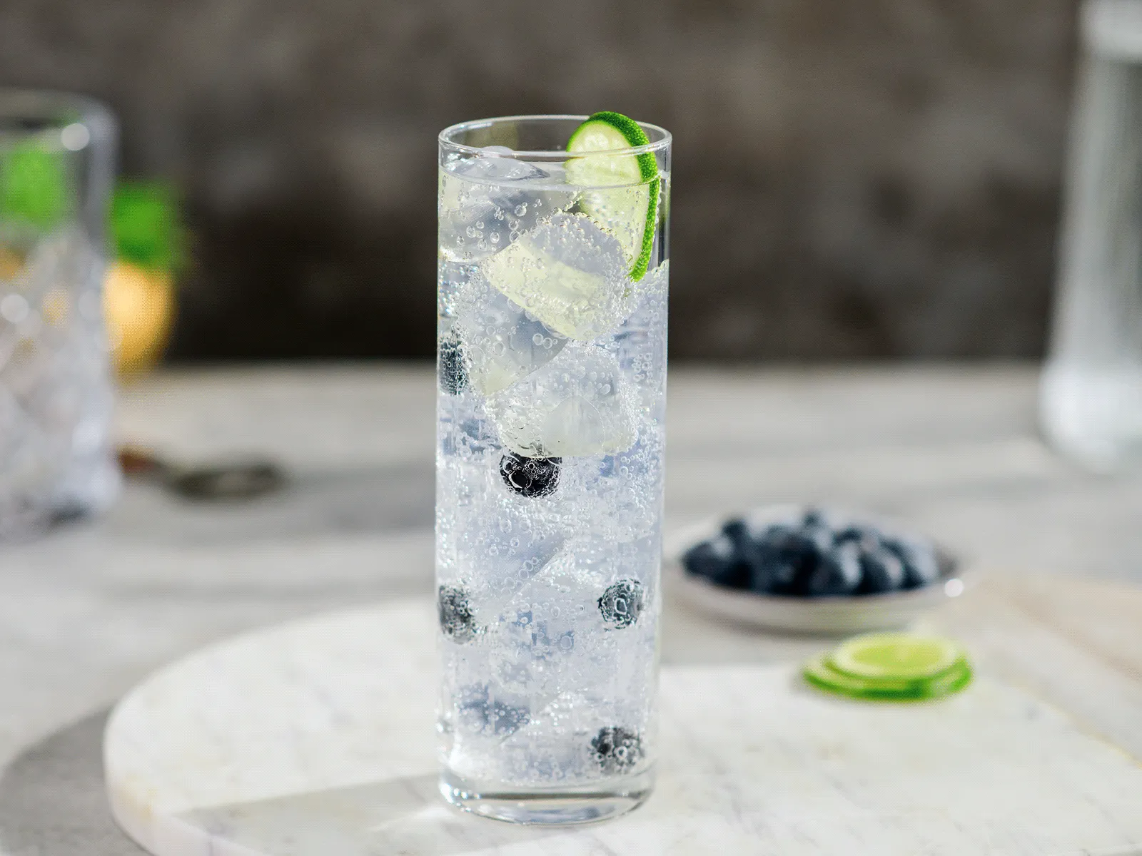A tall glass of sparkling water with ice, blueberries, and a lime wedge garnish.