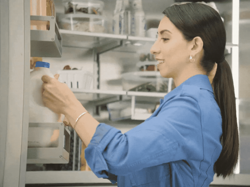 Side profile of a woman taking a carton from a brightly lit refrigerator door shelf.