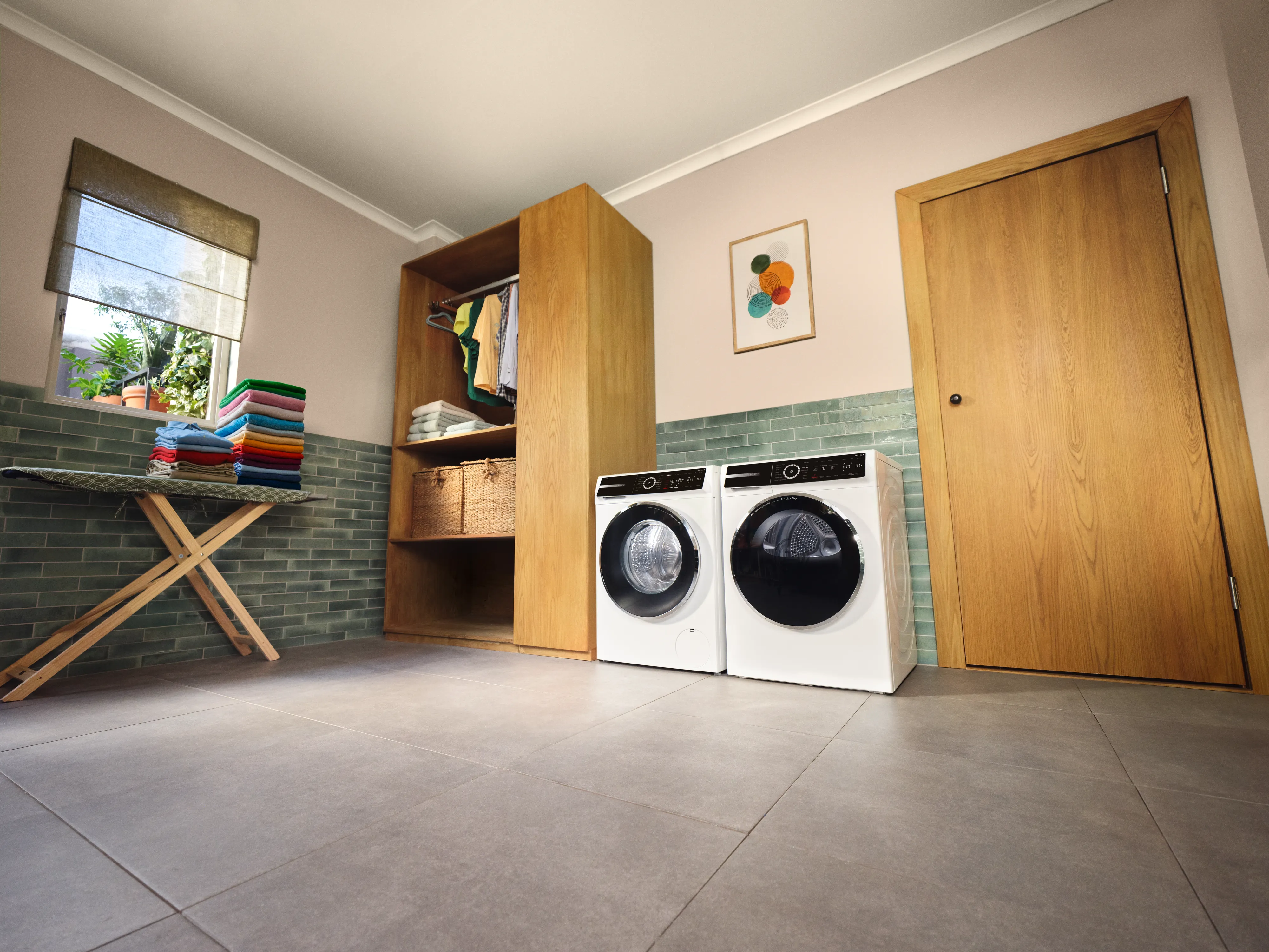 Buanderie avec lave-linge et sèche-linge blancs encastrés sous un meuble en bois, table pliante avec linge plié, mur carrelé vert et porte en bois.