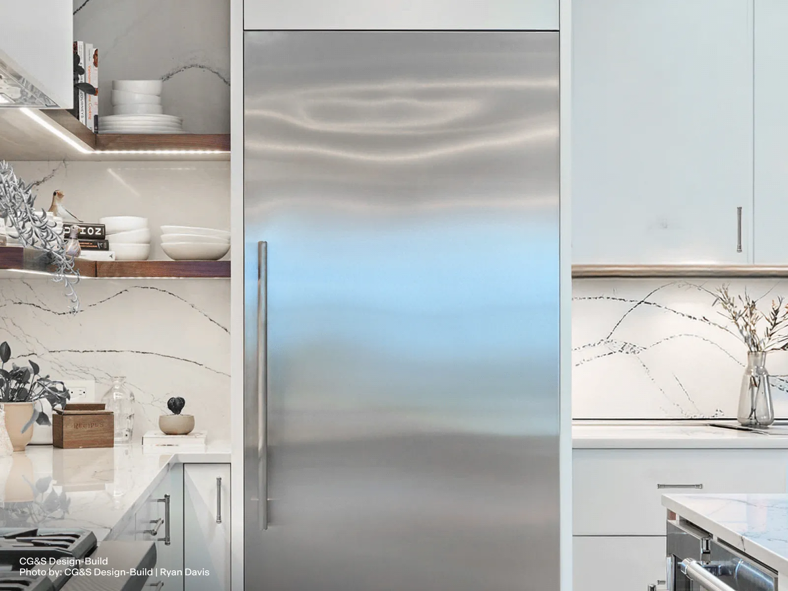 Built-in stainless steel refrigerator column in a modern kitchen, surrounded by white cabinetry, marble backsplash, open shelves with dishes, and countertop accessories.