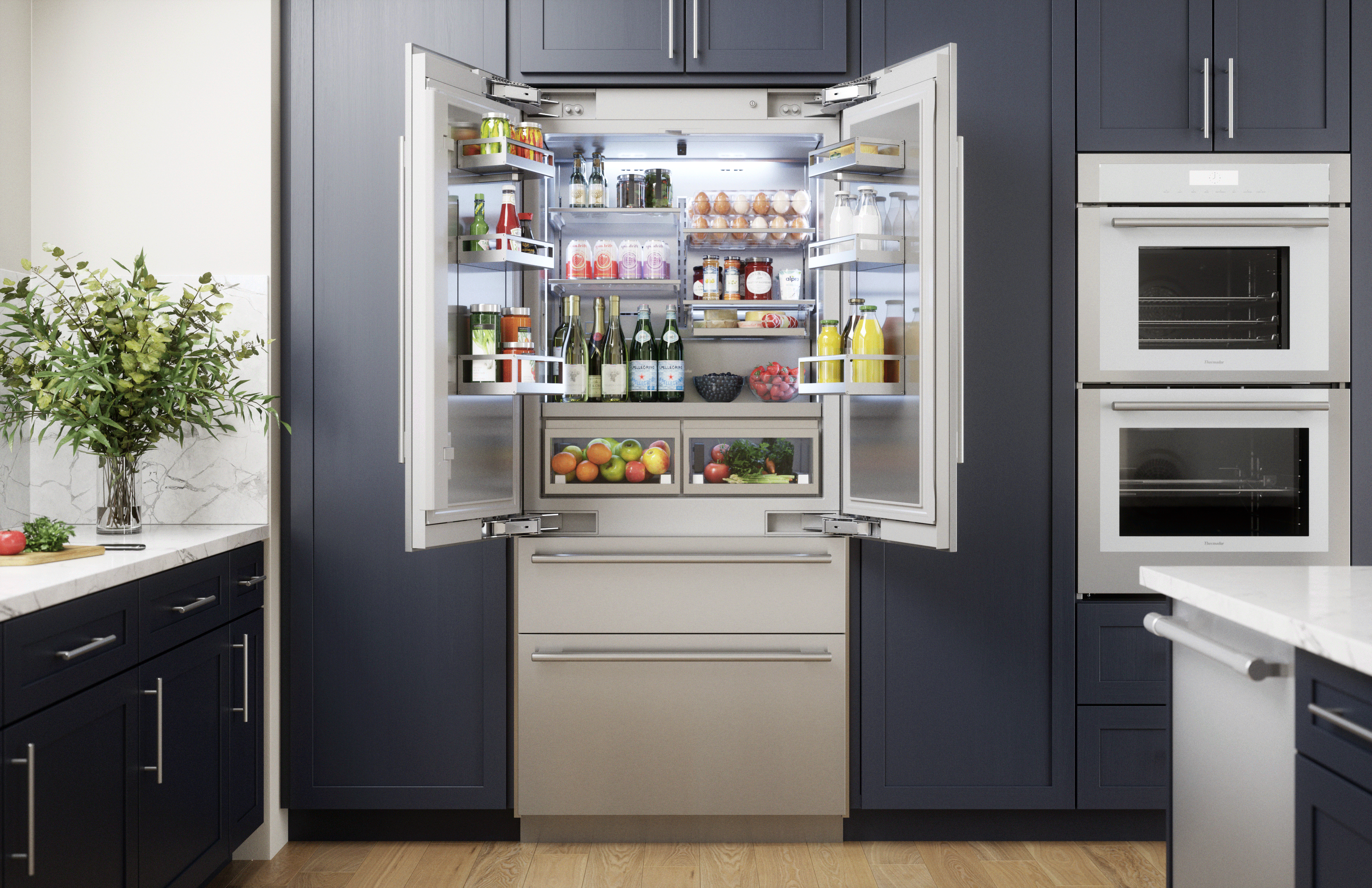 Open built-in French door refrigerator with organized shelves displaying bottles, jars, eggs, and fresh produce, integrated into a modern kitchen with dark cabinetry and stainless steel double ovens nearby.