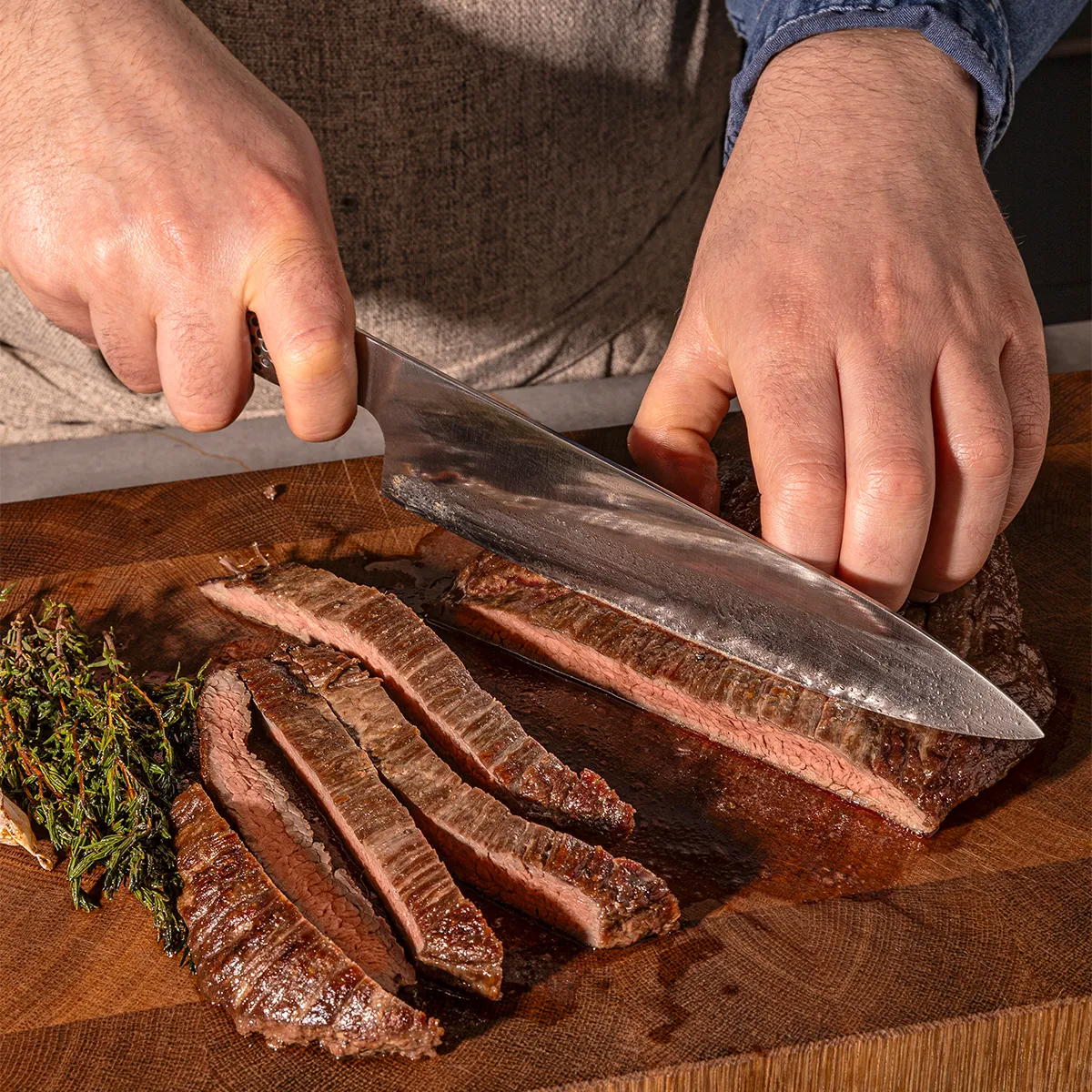 Sliced steak on wooden cutting board, garnished with herbs, knife in hand, preparing a meal.