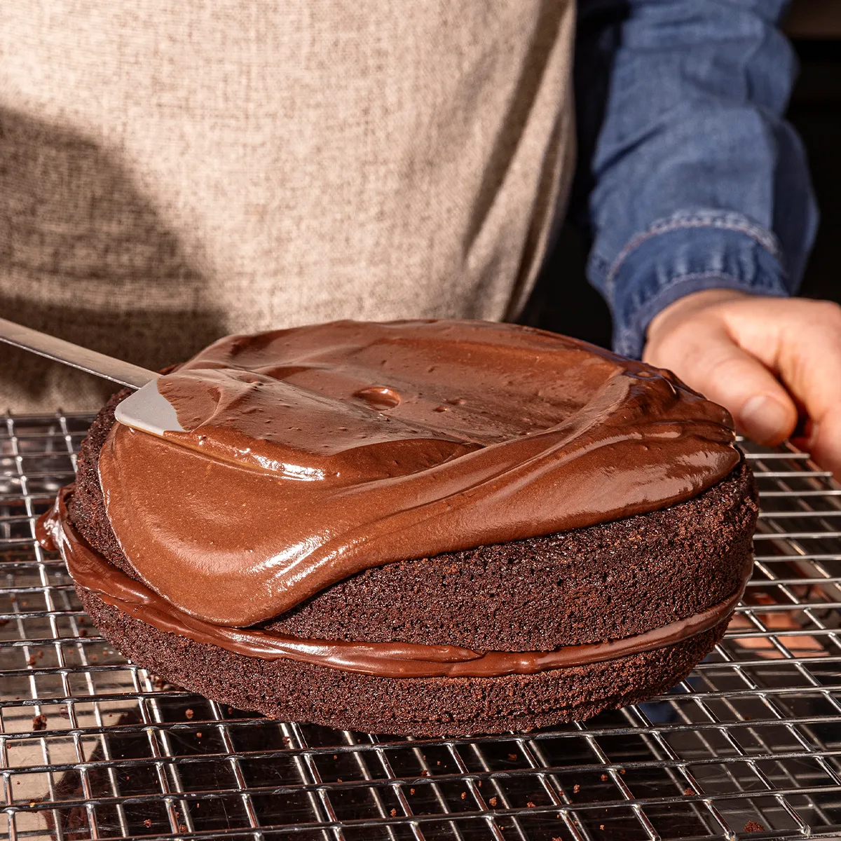 Chocolate cake being frosted on a cooling rack, hands applying chocolate glaze.