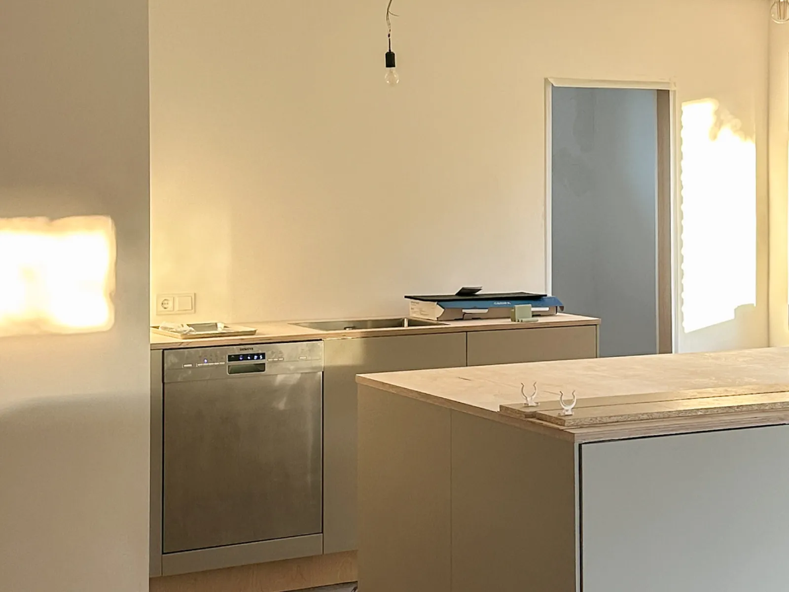 Minimalist kitchen with built-in dishwasher, countertop island with wooden panels, and sunlight streaming through a window.
